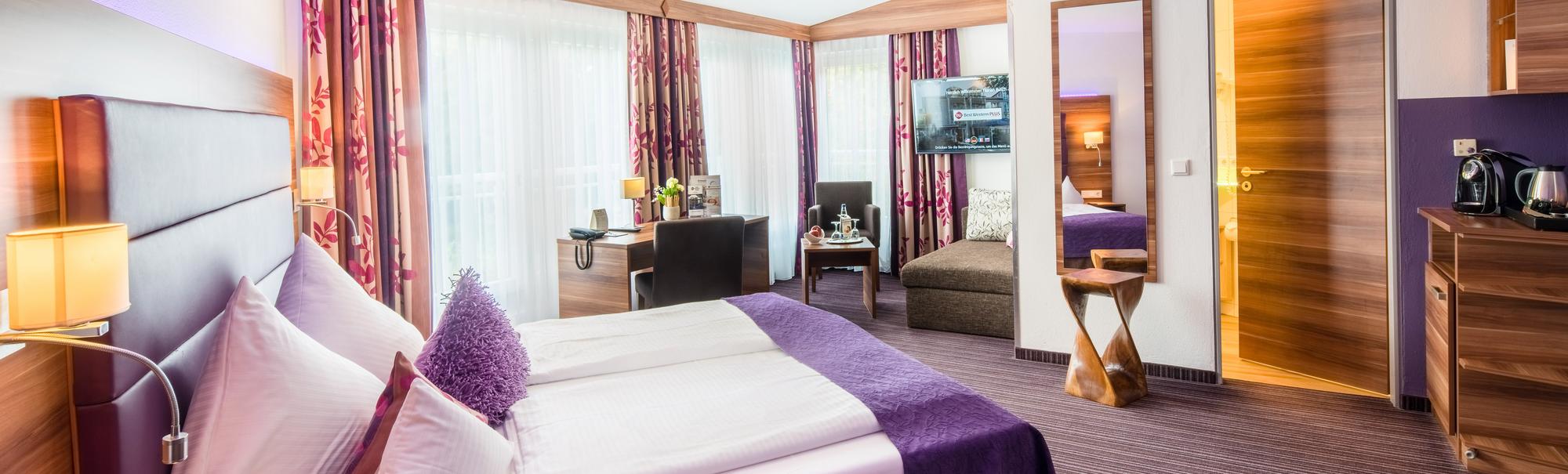 Schlafzimmer mit weiß-lila Bettwäsche, Nachttisch, Wandbildern und Tageslicht durch Fenster.