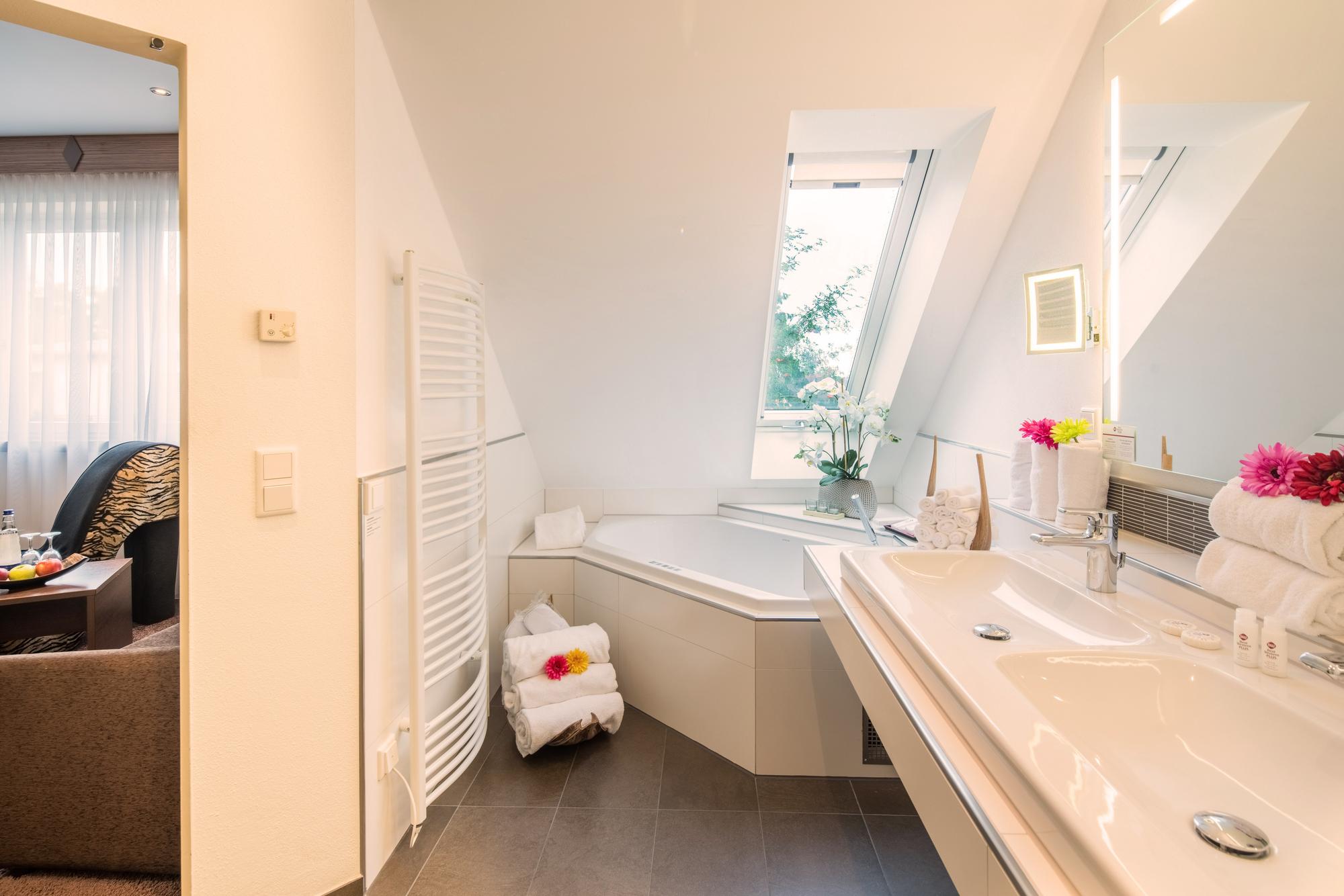 Helles Bad mit Dachschräge, Badewanne, Waschbecken, Tageslichtfenster und offenem Zugang zum Nebenraum.