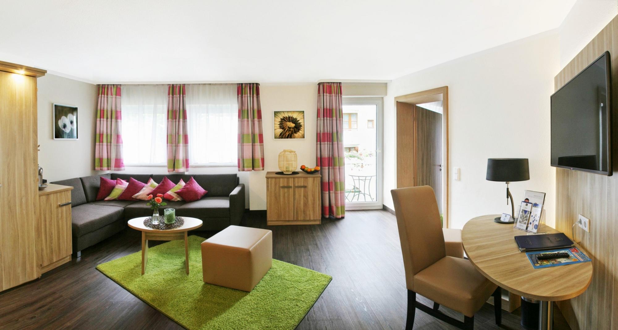 Modernes Wohnzimmer mit schwarzem Sofa, roten Akzenten, grünem Teppich, Schreibtisch und viel Tageslicht.