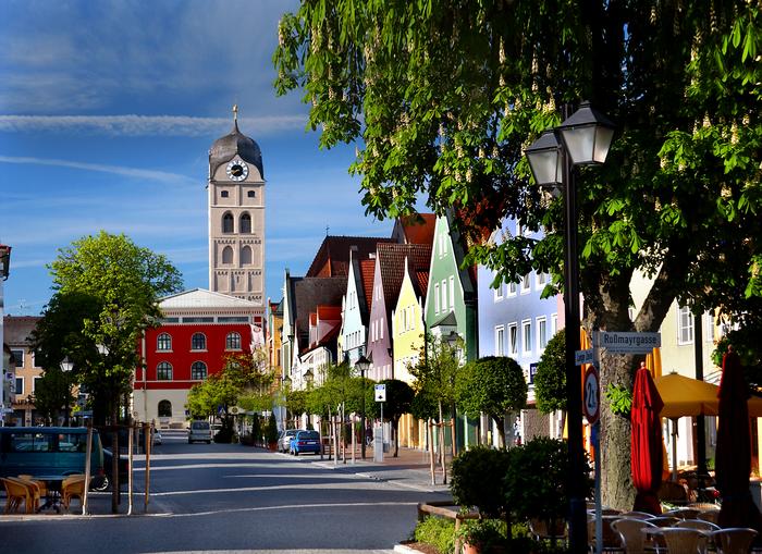 Bunte Häuser und Bäume säumen eine sonnige Straße in europäischer Stadt, im Hintergrund Kirchturm und Schild „Roßmayrgasse“.