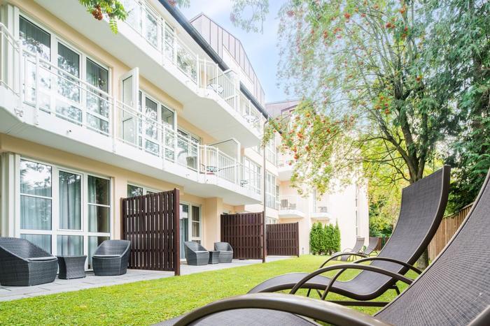 Mehrstöckiges Gebäude mit Balkonen, weiße Fassade, Liegestühle auf Rasenfläche, umgeben von Bäumen und Pflanzen.