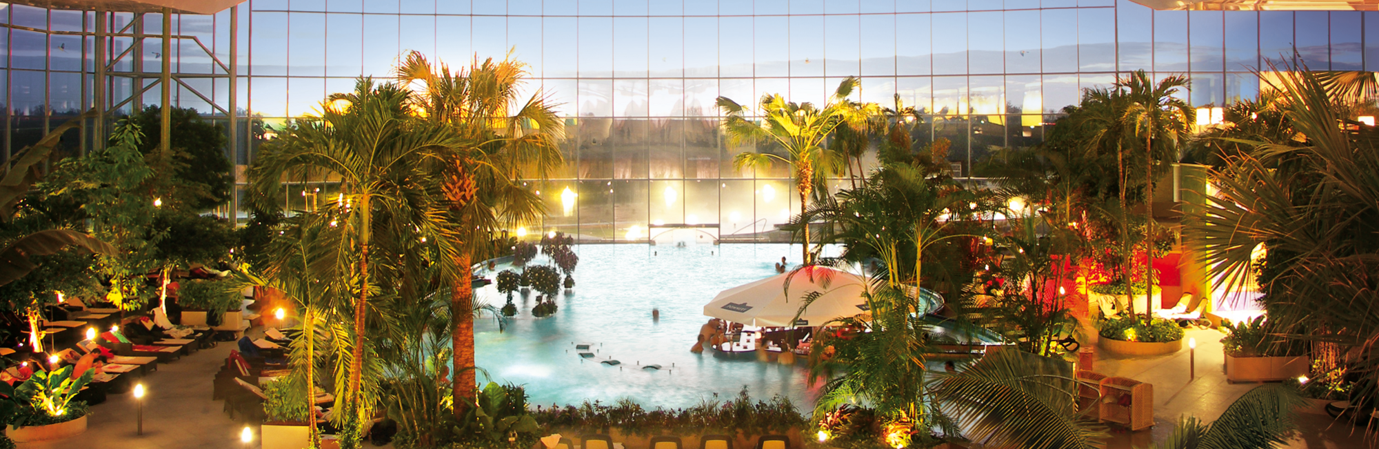 Tropisch gestalteter Indoor-Pool mit Palmen, Liegen, weißem Sonnenschirm und Glaswand für Tageslicht, entspannte Atmosphäre.
