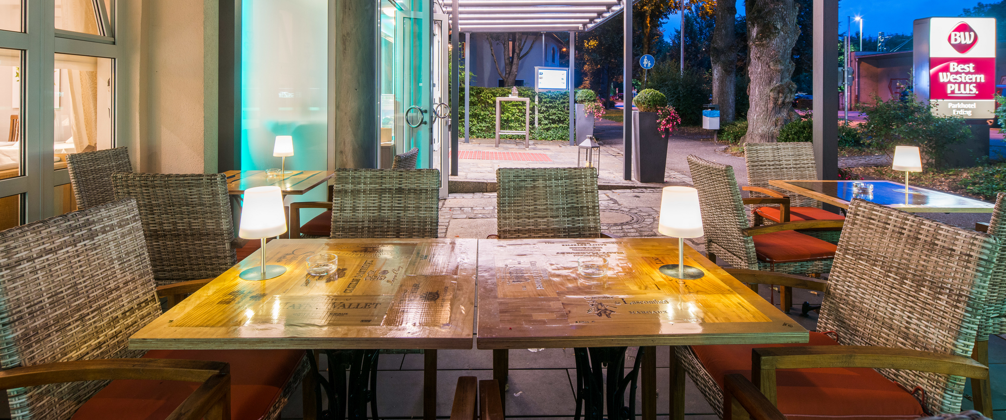 Modernes Restaurant mit Holztischen, gedeckt mit Platzsets und Besteck, gemütliche Beleuchtung und große Fenster mit Tageslicht bei Abendstimmung.