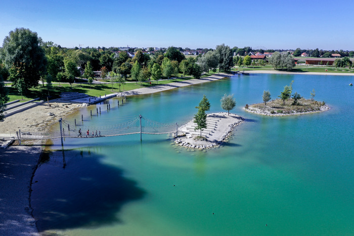Klarer See mit kleinen bewachsenen Inseln, grüner Uferlandschaft und Gebäuden im Hintergrund bei blauem Himmel